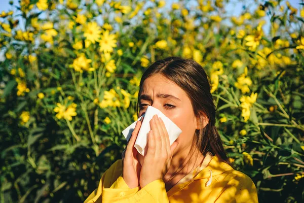 Sneezing young girl with nose wiper among blooming flowers in park. Young woman got nose allergy, flu sneezing nose. The girl suffers from pollen allergy during flowering and uses napkins. Allergy.