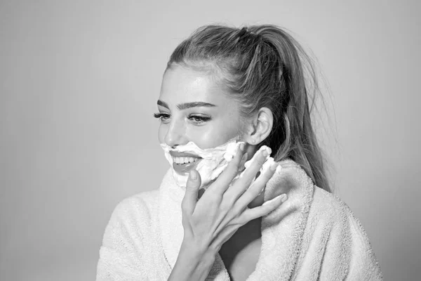 Skin care and shaving concept. Woman with face covered with foam. Lady cares about smooth skin. Girl on smiling face in bathrobe covering face with foam for shaving, grey background.