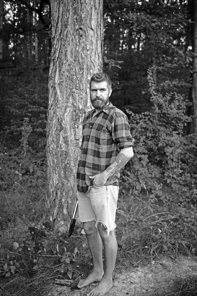Hipster with long beard on natural green landscape. Bearded man with shovel in forest. Tourist in plaid shirt and jean shorts travel barefoot. Traveler hiking on sunny day. Summer vacation concept