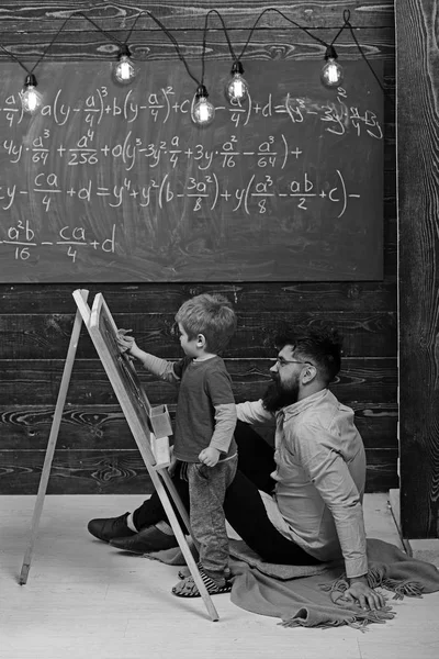 Little genius learning math. Teacher or father helping kid to solve equation on chalkboard. Bearded guy in pink shirt sitting on the floor