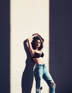 Kadın sakin yüzünde duvara yakın güneşli günde poz. Bayan siyah sütyen ve kot pantolonlu seksi ve çekici görünüyor. Sıcak ve seksi kavramı. Kız esmer çıplak göbek güneş ışığı altında rahatlatıcı ile.