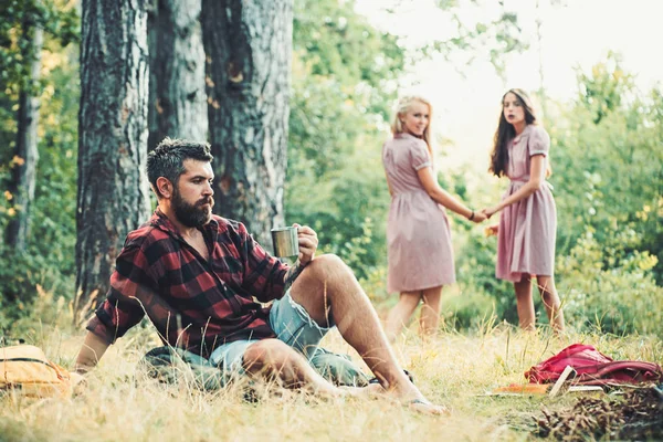 Przyjaciele camping w lesie. Dwie dziewczyny, trzymając się za ręce spojrzeć wstecz na człowiek siedzi na trawie. Widok z boku Brodaty facet siedzi na zielony łąka — Zdjęcie stockowe