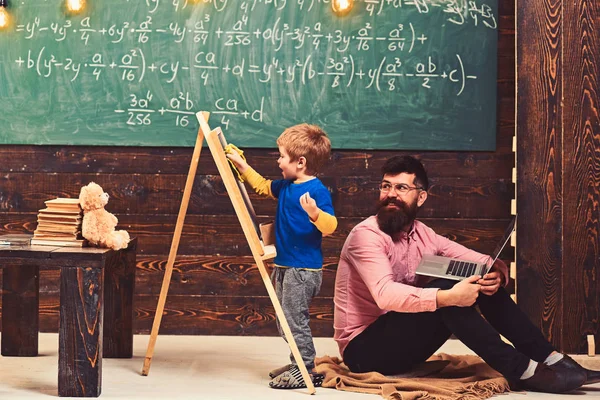 Smiling teacher in glasses looks at kid wiping chalkboard. Father and excited blond kid learning math. Side view boy and man sitting on floor with laptop