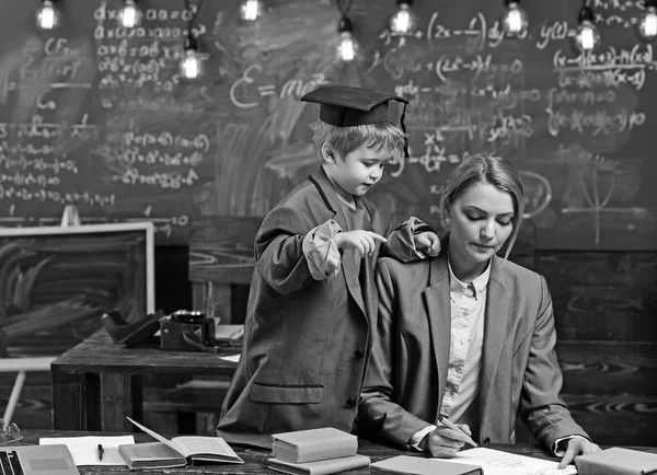 Son and mother have school lesson. Genius boy help woman write homework at desk. Little child in big suit coat and graduation cap teach woman. Back to school. Teaching and learning concept — Stock Photo, Image