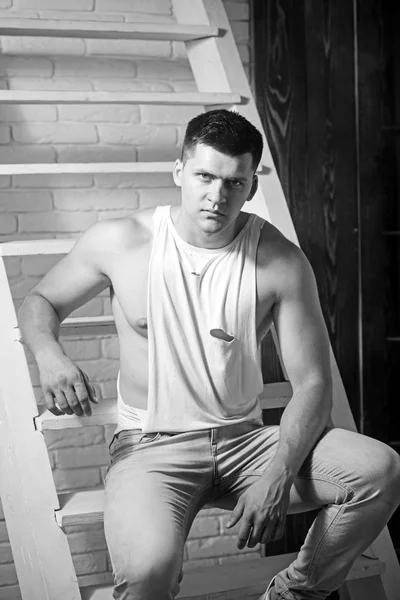 Athletic macho with muscular chest and hands. Young man sit on ladder. Guy in trendy tank top and jeans fashion. Fashion model with stylish hair. Mens fashion style and trend — Stock Photo, Image