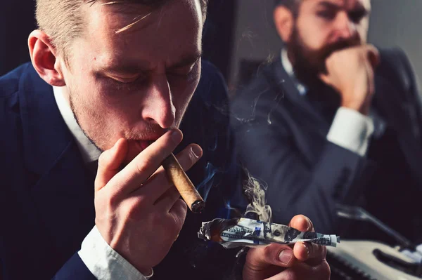 He has megabucks. Businessmen have money to burn. Man light up cigar from money banknote. Rich man smoking during business meeting. Business partners writing financial report. Waste of money