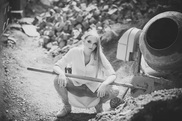 Beauty model s make-upem na cement mixer. Žena pracovní košili a džínách na staveniště, módní. Žena má dlouhé blond vlasy, účes drží lopatu. Móda dívka práce ve stavebnictví, vogue — Stock fotografie