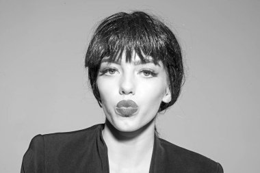 Yüz öpüşmeyle kız siyah resmi ceket giyer. Siyah peruk Bayan gri arka plan üzerinde olun. Öpücük ve ruj kavramı. Kadın çekici kırmızı dudaklar ile kameraya görünüyor.