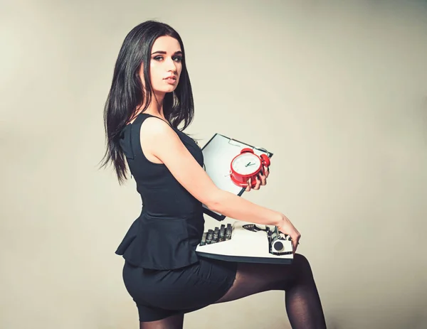 Seductive secretary holding clipboard and typewriter in her hands. Side view beautiful brunette standing with her leg bent isolated on gray background