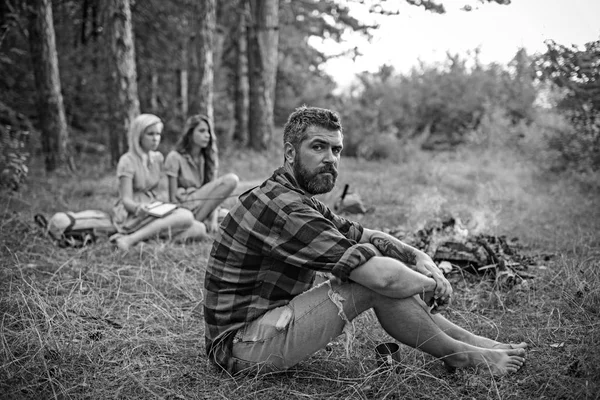 Bearded man relax at bonfire with women on blurred background. Hipster with beard sit at campfire. Friends enjoy camping in forest. Summer vacation concept. Camping, hiking and travel