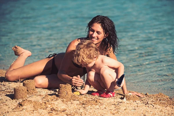 Маленький мальчик строит песочный замок у активной матери, вдоль берега воды на пляже. Ребенок так гордится, что может идти в ногу со своей матерью — стоковое фото