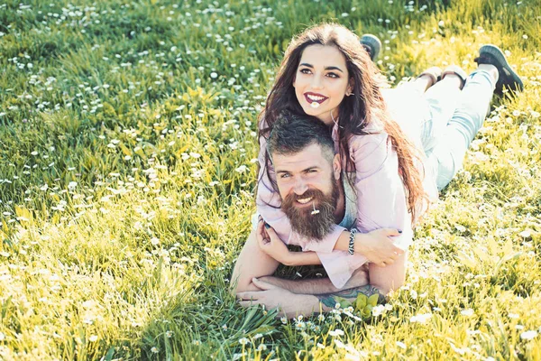 Man and woman laying on grass spring day. Spring leisure concept. Couple on happy faces laying at meadow with daisy flowers in mouth, nature on background. Couple in love spend time outdoors and hugs.