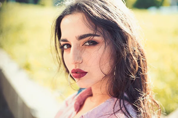 Woman on calm face with make up, nature on background, defocused. Girl with long hair wears unbuttoned shirt on sunny day. Lady looks attractive, close up. Attraction concept.