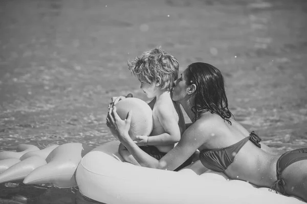 Motherhood concept. Mother lie on mattress near son, kisses and hugs him. Mom and kid spend time together on vacation. Cute child boy with sits on air mattress pineapple shaped, in ocean, sea. — Stock Photo, Image