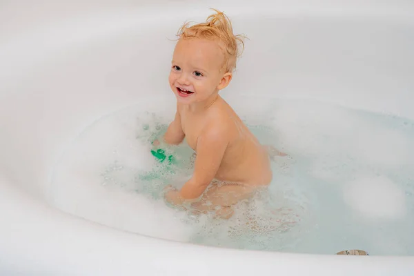 Funny cheerful toddler cleaning body in the bath. Little boy in bathtub with fluffy soap bubble.