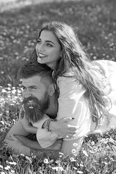 Hombre y mujer acostados en la hierba el día de primavera. Concepto de ocio de primavera. Pareja en caras sonrientes con flores de margarita en la boca, naturaleza en el fondo. Pareja enamorada pasar tiempo al aire libre y abrazos . — Foto de Stock