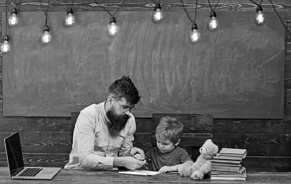 Ученик с учителем в школе. Репетитор помогает ребенку писать письма в блокноте. Мужчина и мальчик сидят за столом с ноутбуком и грудой книг — стоковое фото