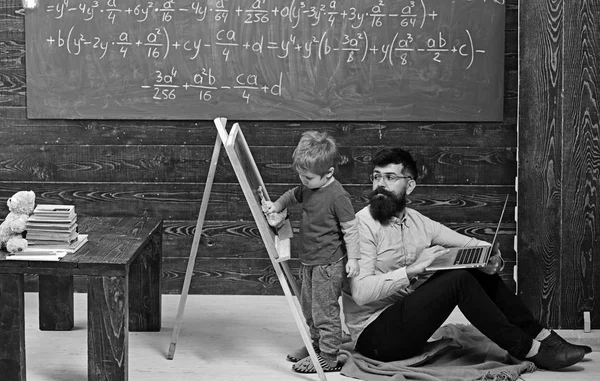 Teacher looking at kid wiping chalkboard. Side view boy and man sitting on floor with laptop. Informal education concept — Stock Photo, Image