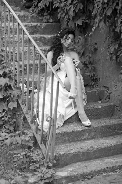 Mode et accessoire de mariage. Femme porter de la dentelle jarretière sur la jambe. Femme sexy en bas lingerie le jour du mariage. Fille avec maquillage nuptial et coiffure. Mariée en robe blanche assis sur les marches en plein air — Photo