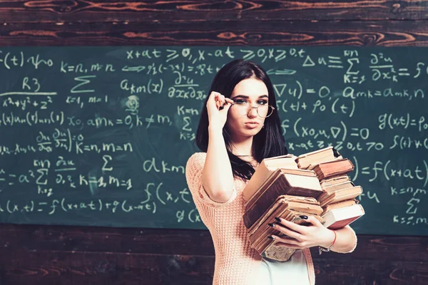 Estudiante morena en cárdigan rosa sosteniendo sus gafas. Hermosa chica universitaria llevando dos montones de libros pesados — Foto de Stock