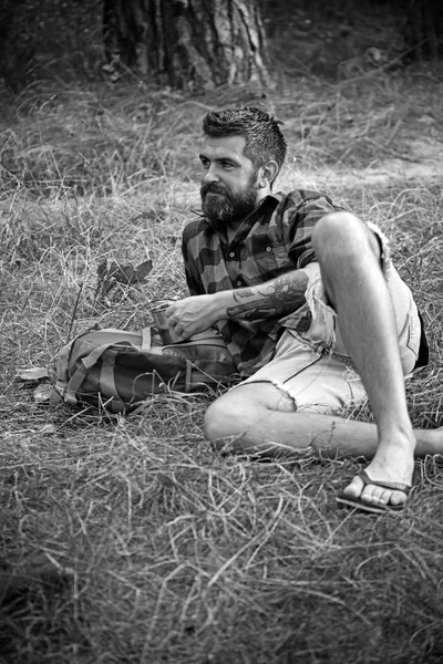 Tourist in plaid shirt hold mug. Hipster with long beard relax on green grass. Bearded man with tea or coffee cup in forest. Camping, drinking and coffee mood. Summer vacation concept — Stock Photo, Image