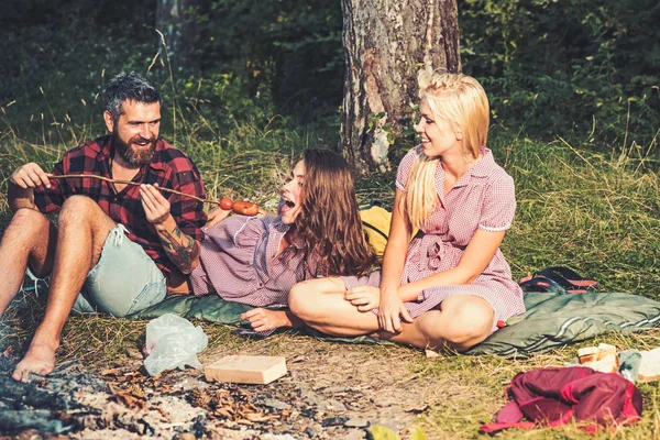 Счастливые друзья, отдыхающие в лесу. Парень кормит свою подружку сосисками фри. Блондинка смотрит на своих улыбающихся друзей — стоковое фото