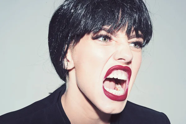 Woman with attractive red lips shouting, close up. Angry boss concept. Girl on scandalous shouting face wears formal jacket. Lady in black wig with make up screaming on grey background.