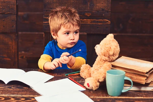 Занятой ребенок в школе. Маленький мальчик сидит за деревянным столом с тетрадями. Блондинка смотрит на своего плюшевого мишку — стоковое фото