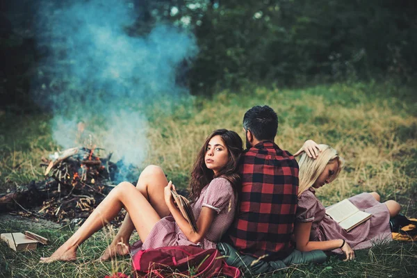 Brunette meisje leunend op haar vriendje tijdens het lezen van boeken. Weer man kijkt bij het kampvuur. Jong koppel in liefde. Vrienden kamperen in bos — Stockfoto