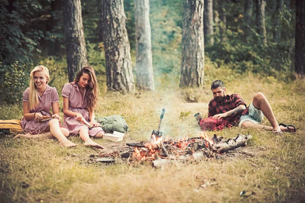 Dziewczyny, siedząc przy ognisku podczas czytania książki. Brodaty facet, leżąc na trawie w lesie. Przyjaciele camping w okresie letnim — Zdjęcie stockowe