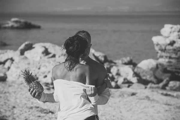 Paar staan op rotsachtige strand en zoenen op zonnige zomerdag. Koppel op vakantie houden ananas. Sexy dame met naakt schouders hugs macho, zee op de achtergrond. Tropische Huwelijksreis concept. — Stockfoto