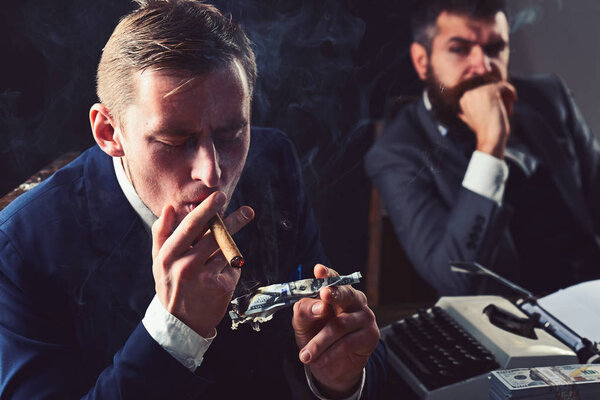 He is lousy with money. Rich man smoking during business meeting. Man light up cigar from money banknote. Businessmen have money to burn. Business partners writing financial report. Waste of money