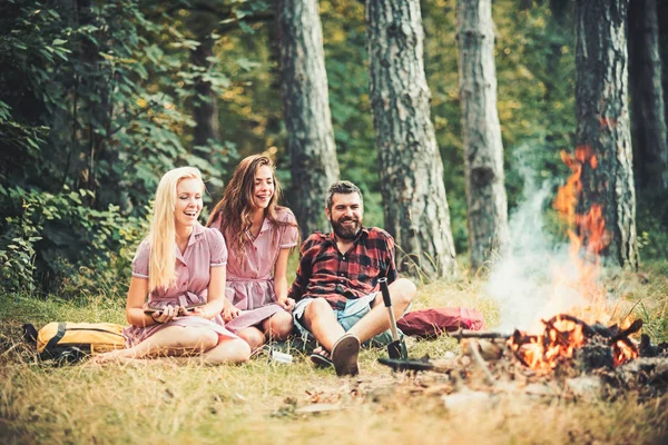 Καλοκαιρινές διακοπές σε κατασκήνωση. Γενειοφόρος άνδρας και γυναίκες χαμόγελο σε φωτιά. Ευτυχής φίλους στο campfire. Hipster στο πουκάμισο palid και τα κορίτσια σε vintage φορέματα Χαλαρώστε στο δάσος. Κάμπινγκ ταξίδια και περιπλάνησης, ρετρό — Φωτογραφία Αρχείου