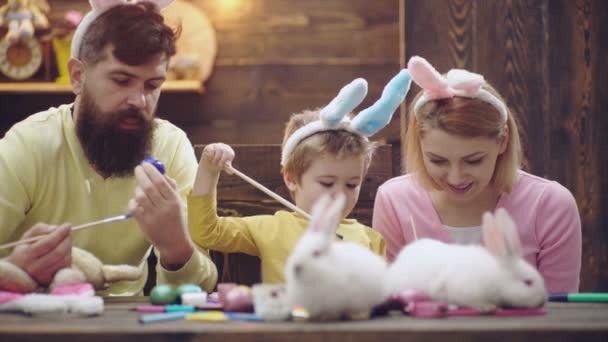 Madre, padre e hijo están pintando huevos. La familia feliz se está preparando para la Pascua. Lindo niño con orejas de conejito. Huevos de Pascua hechos a mano pintados por toda la familia. Huevos sobre fondo de madera . — Vídeos de Stock