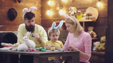 Yumurta boyama güzel çocuk çocuk. Mutlu bir aile Paskalya için hazırlanıyor. Sevimli küçük çocuk çocuk üstünde Paskalya yortusu Bunny kulaklar giyiyor. Anne, baba ve oğul resim yumurta vardır. Ahşap arka plan üstünde Paskalya yortusu.