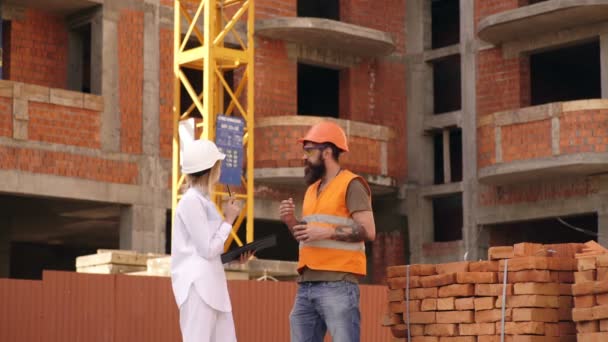 Constructeur en architect gekleed in oranje werk vesten en harde vleermuizen bespreken bouwproces op de open bouwplaats in de buurt van de kraan. Jonge ingenieurs doorlopen van de bouwplaats. — Stockvideo