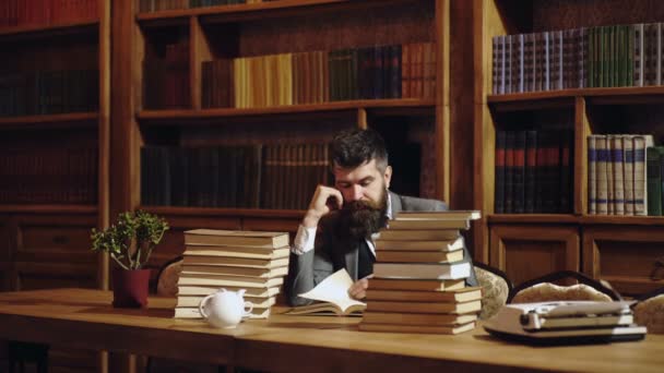 Estudiante masculino serio leyendo un libro en una biblioteca. Hombre con barba leyendo libro sobre fondo de estantería. Hombre en traje clásico, profesor con la cara ocupada concentrada se sienta en la biblioteca cerca de montones de libros . — Vídeos de Stock