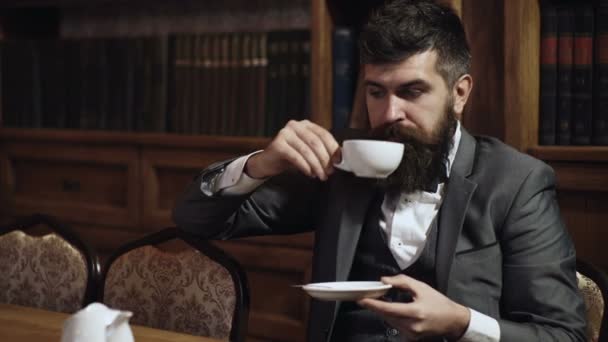 可敬的人在书柜的背景上喝茶。老式的男人拿着杯子和茶。穿着经典西服的男人坐在复古的内部, 图书馆, 书架上的背景. — 图库视频影像
