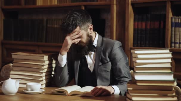 Man in classic suit, professor with concentrated busy face sits in library near piles of books, bookshelves on background. Man or bearded teacher reads book. Research and studying concept. — Stock Video