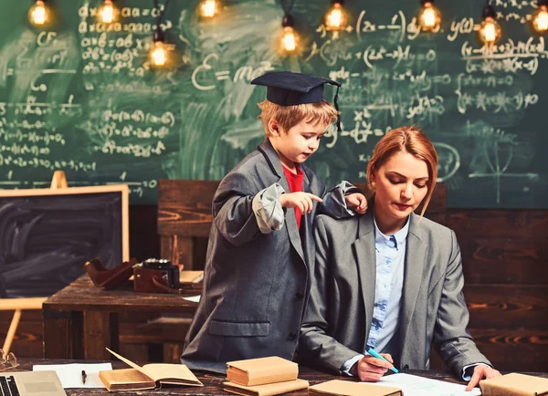 Hijo y madre tienen clase de escuela. Un genio ayuda a una mujer a escribir los deberes en el escritorio. Niño pequeño con abrigo de traje grande y gorra de graduación enseñar a la mujer. De vuelta a la escuela. Concepto de enseñanza y aprendizaje — Foto de Stock