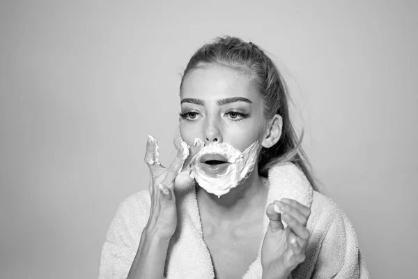 Girl on busy face in bathrobe covering face with foam for shaving, grey background. Woman with face covered with foam. Lady cares about smooth skin. Skin care and shaving concept.