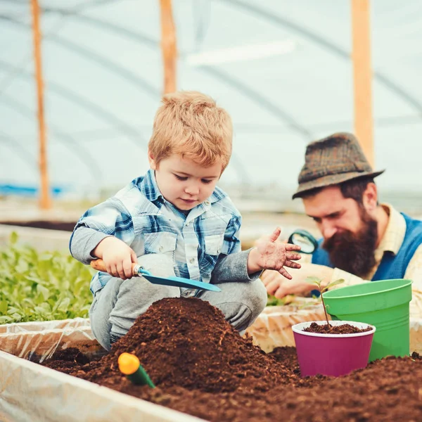 Joli gamin blond assis dans une boîte pleine de terre. Petit garçon jouant pique de jardinage et faisant monticule de terre. Homme à longue barbe et moustache au chapeau debout avec loupe floue — Photo