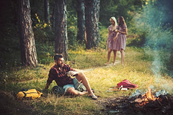 Camping in forest. Man looking at fire while drinking tea. Guy sitting on grass on sunny meadow. Two barefoot girls walking on path.