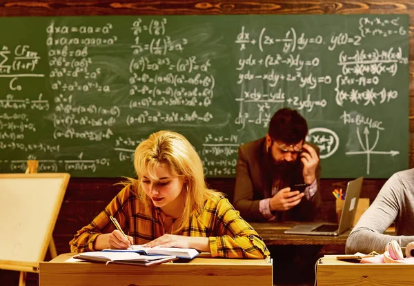 Insegnante in occhiali che legge rapporti di studenti di talento che correggono errori. Studenti attenti che scrivono qualcosa nei loro blocchi appunti mentre sono seduti alle scrivanie in classe . — Foto Stock