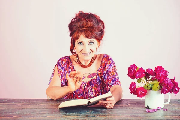 Oude vrouw lezen boek met glazen op bloemen. — Stockfoto