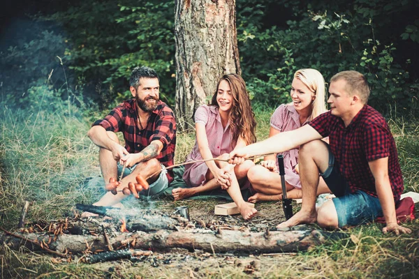 Молодые пары устраивают пикник в лесу. Бородач и его лучший друг жарят сосиски над огнем. Счастливые люди, сидящие у костра, дружба и концепция отдыха — стоковое фото