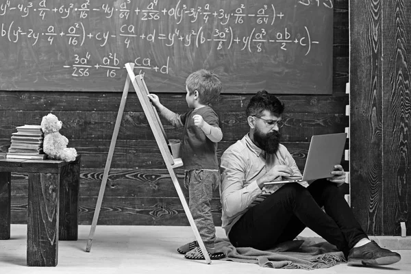 Aula de aritmética na escola. Criança escrevendo no quadro enquanto professor concentrado trabalha no laptop. Vista lateral sentado homem e criança de pé de volta para trás — Fotografia de Stock