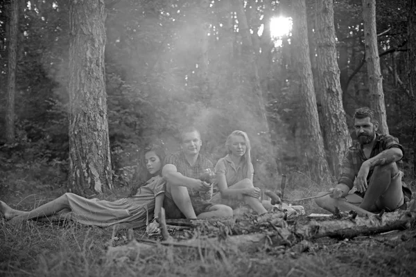 Gruppo di giovani amici che si accampano nella foresta. Due coppie che si godono un picnic nella calda serata estiva. Stanco uomo barbuto cucinare cibo sopra il falò — Foto Stock