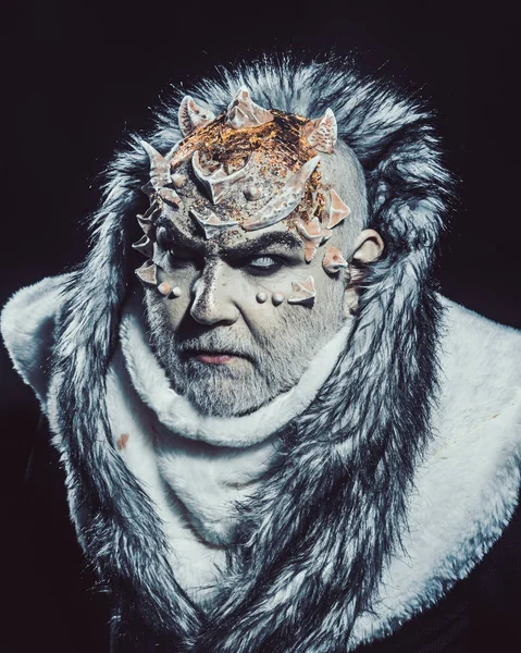 Senior man with white beard dressed like monster. Alien, demon, sorcerer makeup. Dark arts concept. Demon on black background, close up. Man with thorns or warts in fur coat