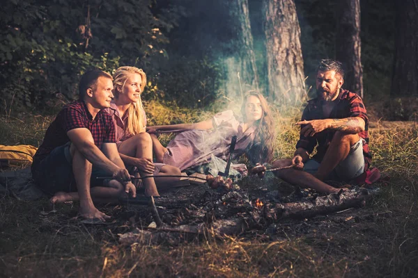 Grupp av vänner camping i skogen. Två par steka korv på pinnar. Ungdomar talar runt lägerelden — Stockfoto